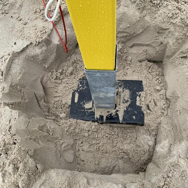 Nahaufnahme eines gelben Pfostens in einem Sandfundament mit nassen Sandkörnern und einer schwarzen Metallplatte.