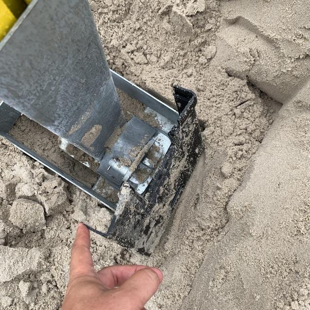 Nahaufnahme eines metallischen Werkzeugs, das in sandigem Boden steckt, während eine Hand darauf zeigt.