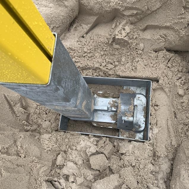 Metallrahmen mit gelbem Pfosten ist im Sand verankert, umgeben von losem Sandmaterial.