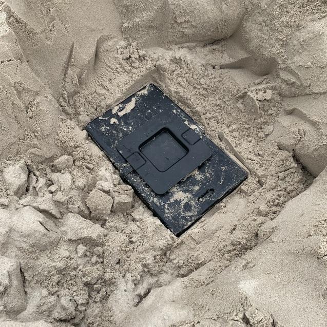 Schwarzes Objekt teilweise im Sand vergraben, umgeben von feinem, hellem Sand.