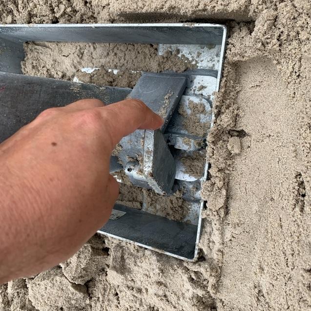 Hand, die auf einen Metallwinkel zeigt, der in einen Haufen Sand im Hintergrund eingebettet ist.