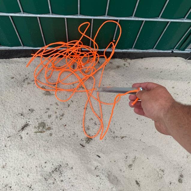 Ein verwickeltes orangenes Seil liegt auf sandigem Untergrund, während eine Hand mit einer Schere daneben gehalten wird.