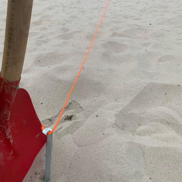 Nahaufnahme eines roten Schaufelblatts neben einem Metallsteg und einer leuchtend orangefarbenen Schnur auf Sand.