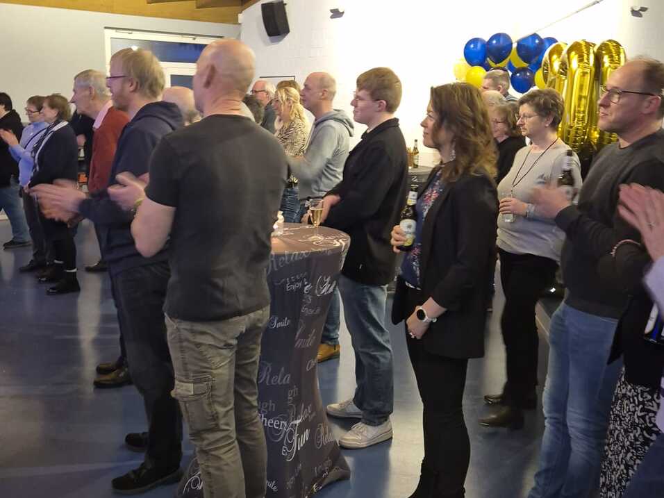 Viele Personen stehen in einem Raum mit blauem Fu&szlig;boden und h&ouml;ren einem Vortrag zu.