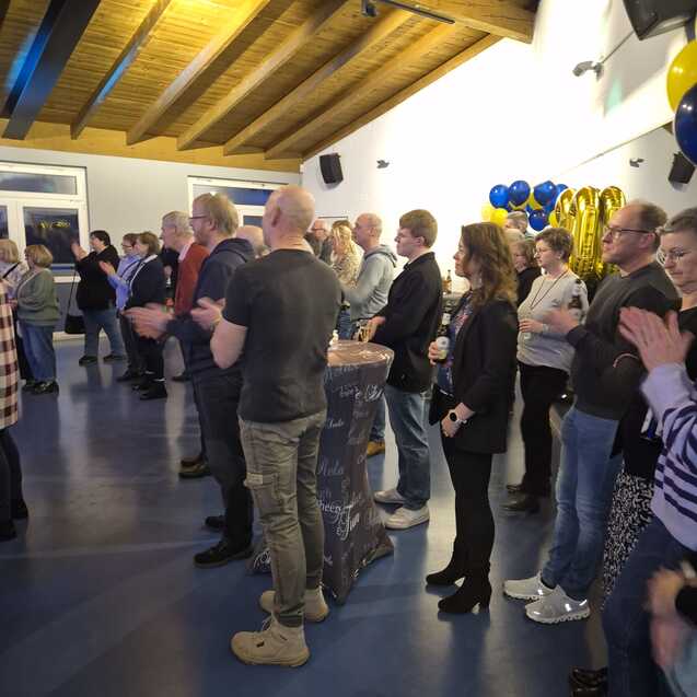 Viele Personen stehen in einem Raum mit blauem Fu&szlig;boden und h&ouml;ren einem Vortrag zu.