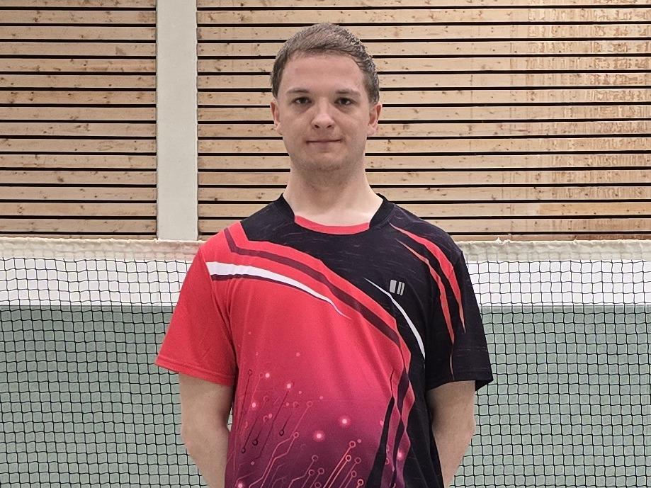 Ein Badmintonspieler in rotem Trikot steht auf einem Court, im Hintergrund ist das Netz und einige Federb&auml;lle verteilt.