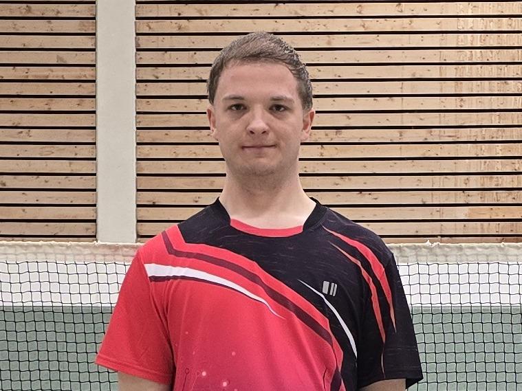Ein Badmintonspieler in rotem Trikot steht auf einem Court, im Hintergrund ist das Netz und einige Federb&auml;lle verteilt.