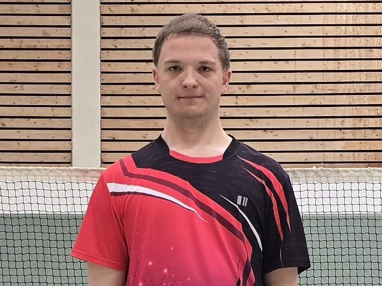 Ein Badmintonspieler in rotem Trikot steht auf einem Court, im Hintergrund ist das Netz und einige Federb&auml;lle verteilt.