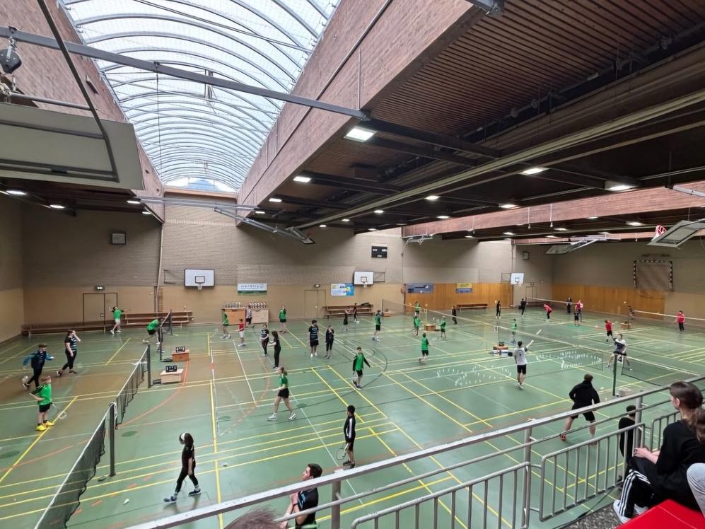 Gro&szlig;e Sporthalle mit mehreren Badmintonspielern in gr&uuml;nen Trikots, Zuschauer sitzen auf der Trib&uuml;ne.