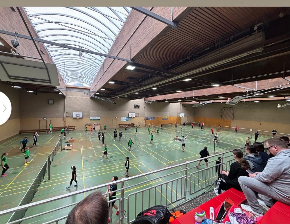 Gro&szlig;e Sporthalle mit mehreren Badmintonspielern in gr&uuml;nen Trikots, Zuschauer sitzen auf der Trib&uuml;ne.