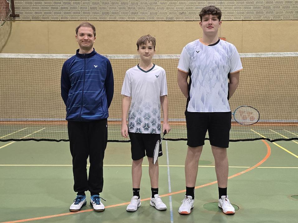 Ein Trainer und zwei Spieler stehen vor einem Badmintonnetz in einer Sporthalle mit grünem Fußboden.