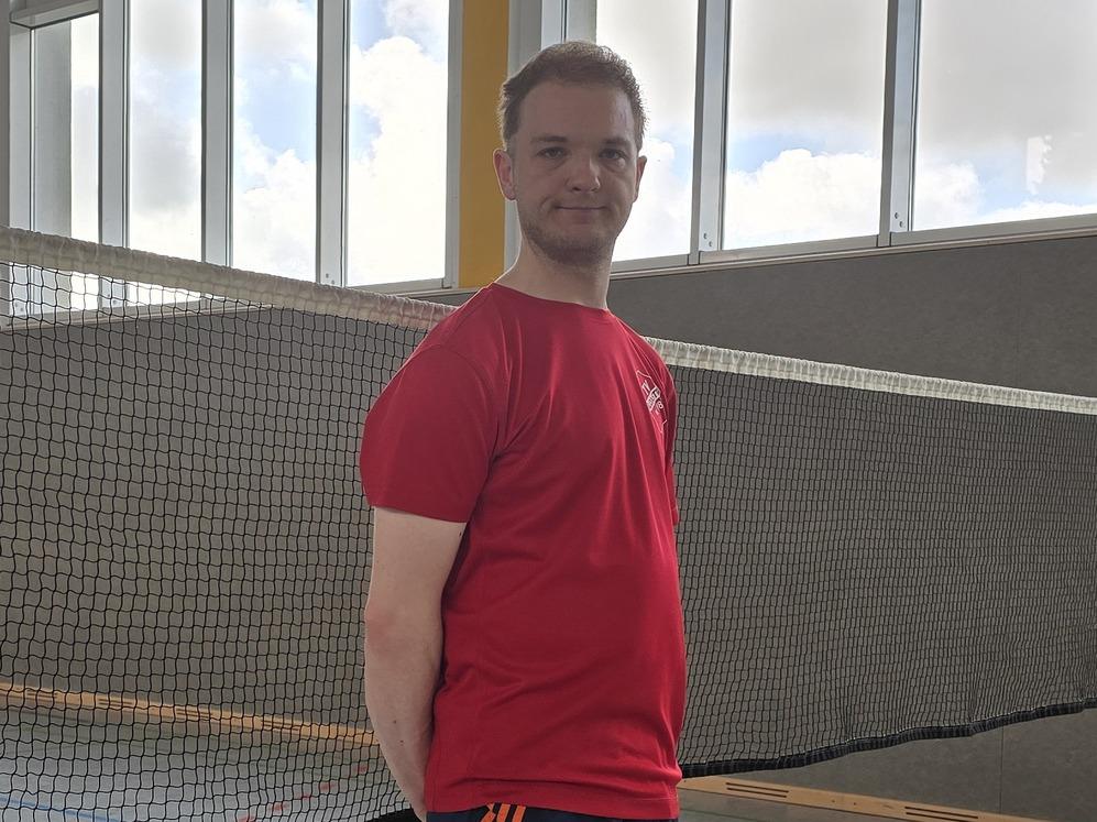 Ein Mann steht vor einem Badmintonnetz in einer Halle, bereit f&uuml;r ein Spiel auf dem Indoorplatz.