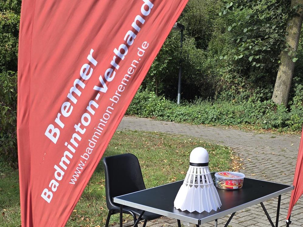 Ein Tisch mit einem &uuml;bergro&szlig;en Badmintonball und einer Schale steht neben zwei St&uuml;hlen auf Pflastersteinen, links zwei rote Beachflags mit der Aufschrift 'Bremer Badminton-Verband' und einer Webadresse.
