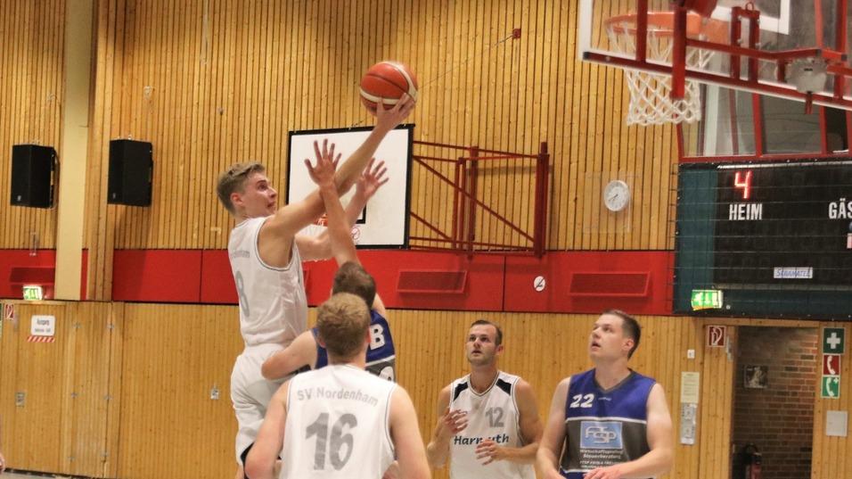 Spielszene eines Basketballspiels, Spieler im weißen Trikot steigt zum Korb, Zuschauer und Tafel im Hintergrund.