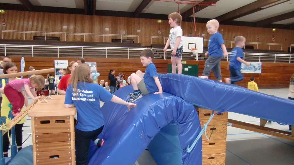 Kinder spielen auf einem Bewegungs parcours mit einem blauen Schaumstoffelement und Holztisch, im Hintergrund eine Sporthalle.