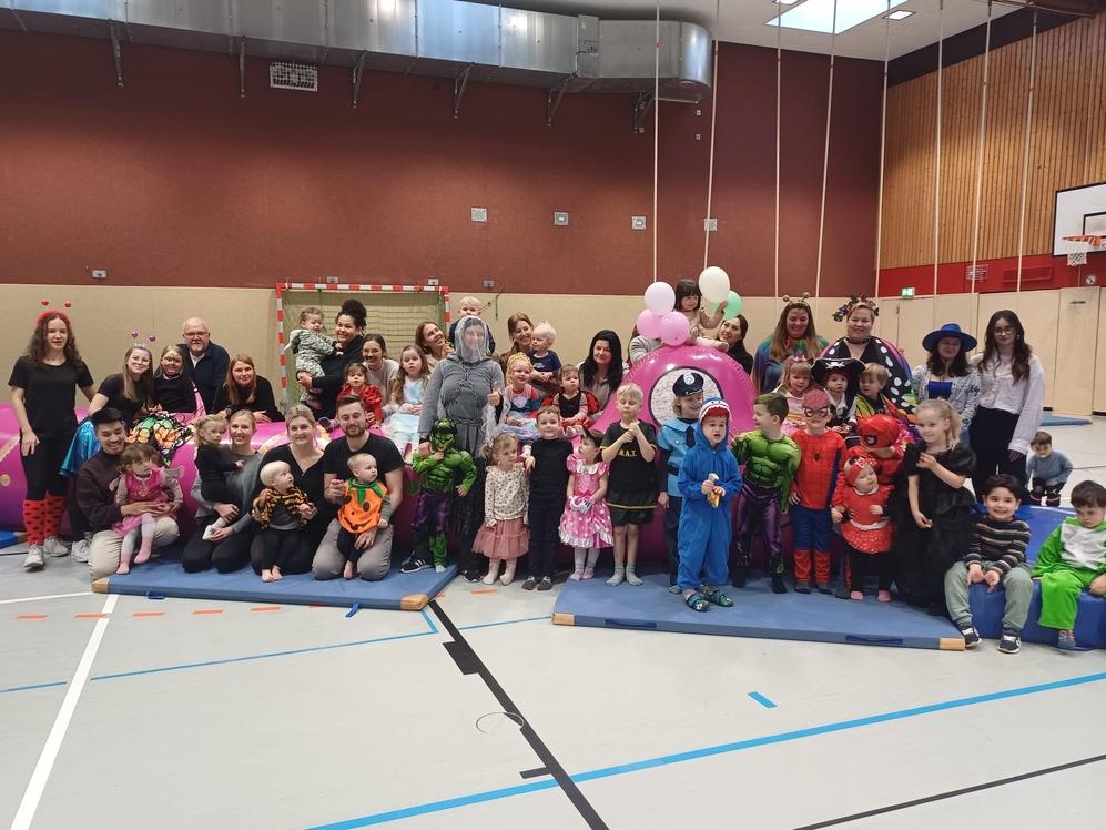 Gruppenfoto von Kindern und Erwachsenen in bunten Kost&uuml;men in einer Sporthalle beim Feiern. Luftballons im Hintergrund.