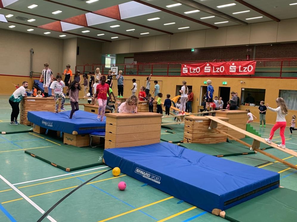 Kinder springen auf einem Trampolin und klettern über Kästen und große blaue Matten auf dem Gymnastikboden.