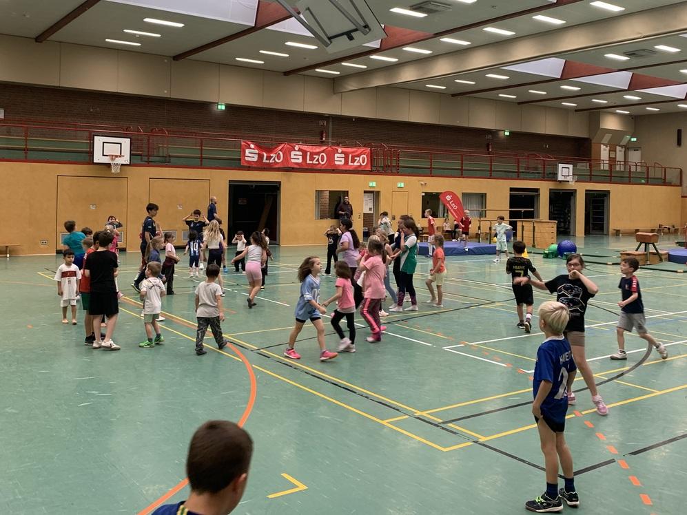 Kinder spielen und bewegen sich in einer gro&szlig;en Sporthalle mit Basketballk&ouml;rben und einer Trib&uuml;ne im Hintergrund.