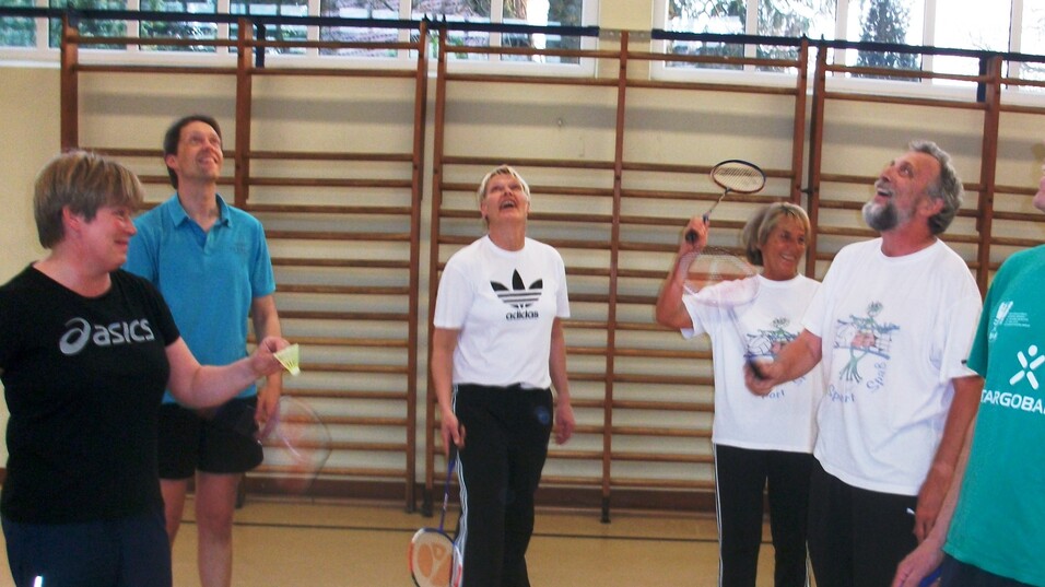 Sechs Personen mit Badmintonschlägern in einer Sporthalle, lachen und konzentrieren sich auf einen Federball.
