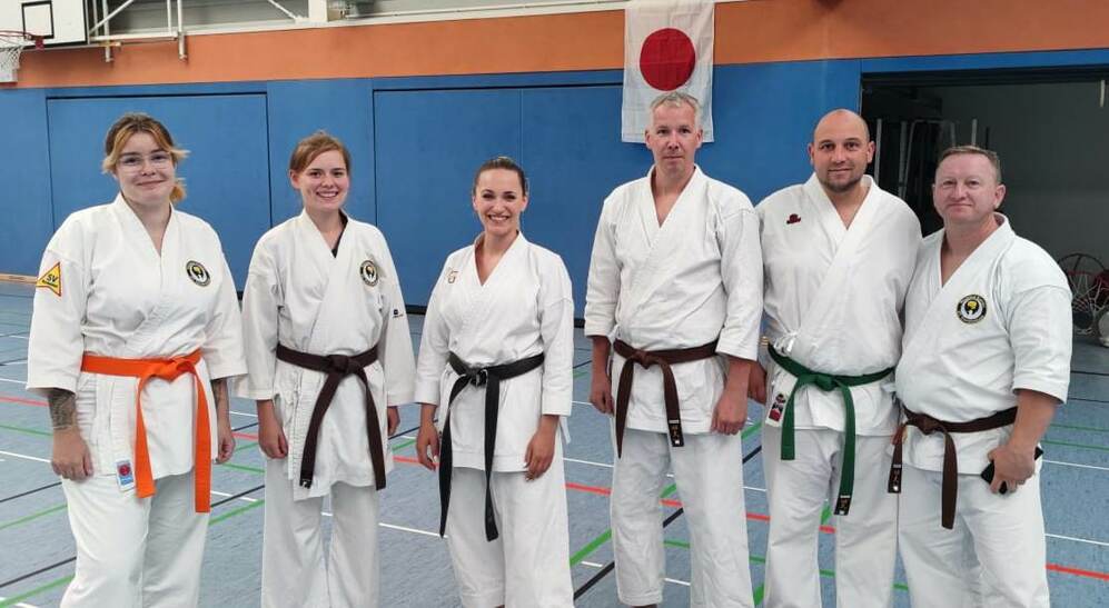 Sechs Karateka in wei&szlig;en Trainingsanz&uuml;gen und unterschiedlichen G&uuml;rtelfarben posieren in einer Sporthalle.