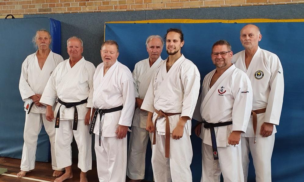 Gruppenfoto von sieben Karateka in wei&szlig;en und braunen Gi, stehend vor einer blauen Wand in einer Sporthalle.