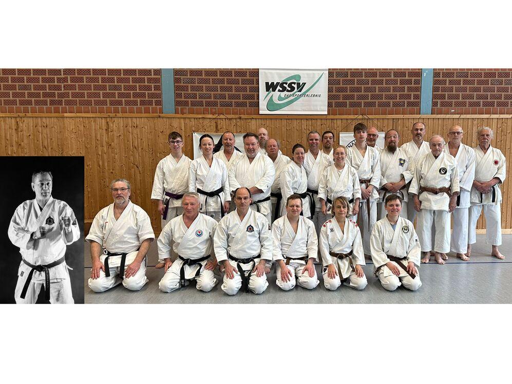 Gruppe von Erwachsenen in wei&szlig;en Karateanz&uuml;gen mit schwarzen G&uuml;rteln in einer Sporthalle vor einer Wand mit Holzverkleidung und einem Banner mit dem Schriftzug 'WSSV'.