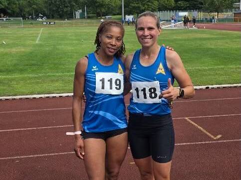 Zwei Sportlerinnen in blauen Trikots mit Startnummern stehen auf einer Laufbahn und l&auml;cheln in die Kamera.