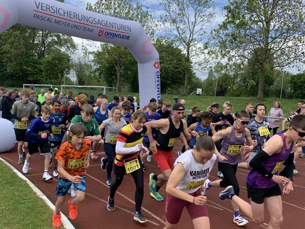 Gruppe von Läuferinnen und Läufern beim Start auf einer Laufbahn unter einem aufblasbaren Torbogen mit Werbeaufschrift.