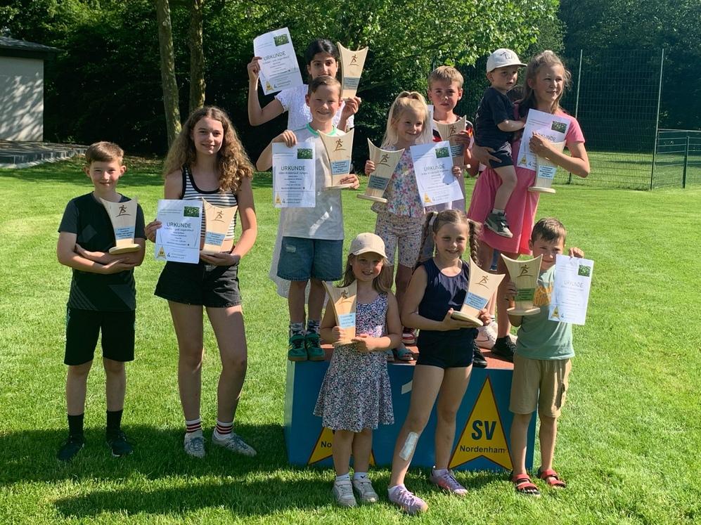 Kinder und Jugendliche stehen mit Urkunden auf und vor einem Siegerpodest auf einer Wiese.
