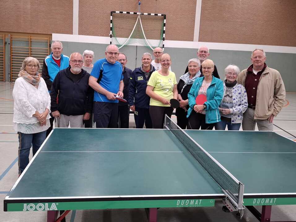Gruppe älterer Menschen steht in Sportkleidung hinter einer Tischtennisplatte in einer Turnhalle.
