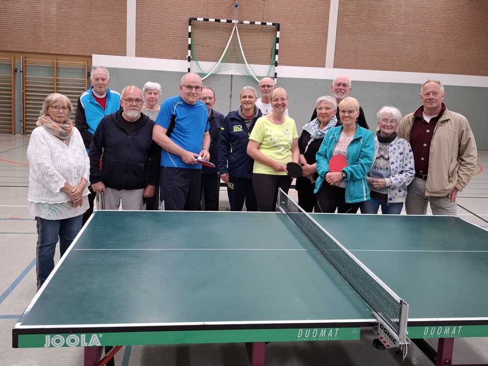 Gruppe &auml;lterer Menschen steht in Sportkleidung hinter einer Tischtennisplatte in einer Turnhalle.