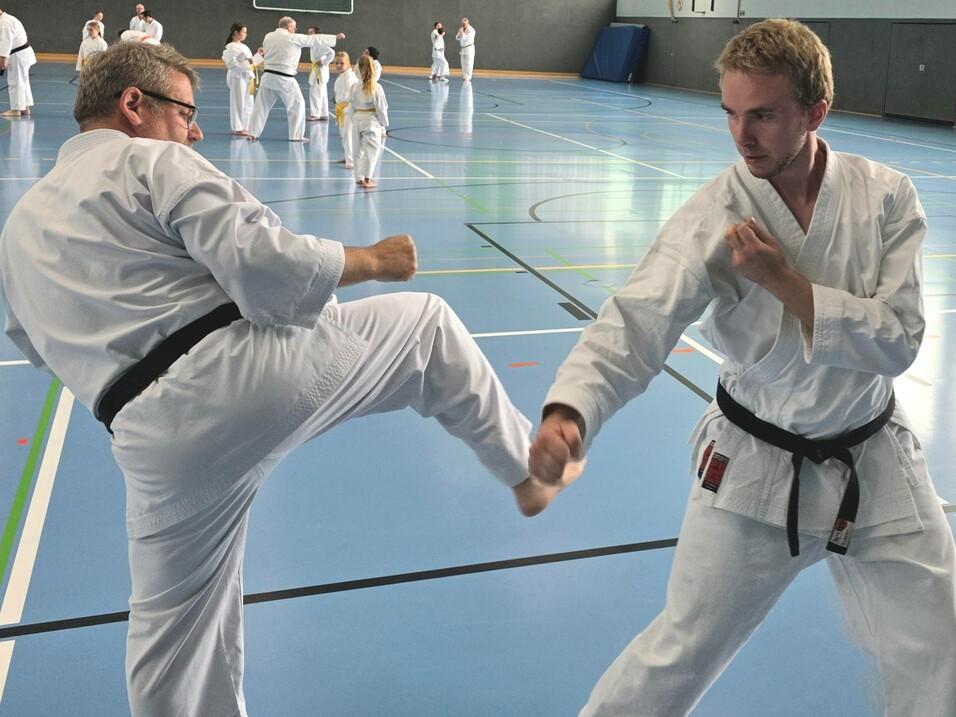 2 Sportler in weißen Anzügen kämpfen in einer Sporthalle mit blauem Fußboden. Im Hintergrund sind noch weitere Aktive zu sehen.