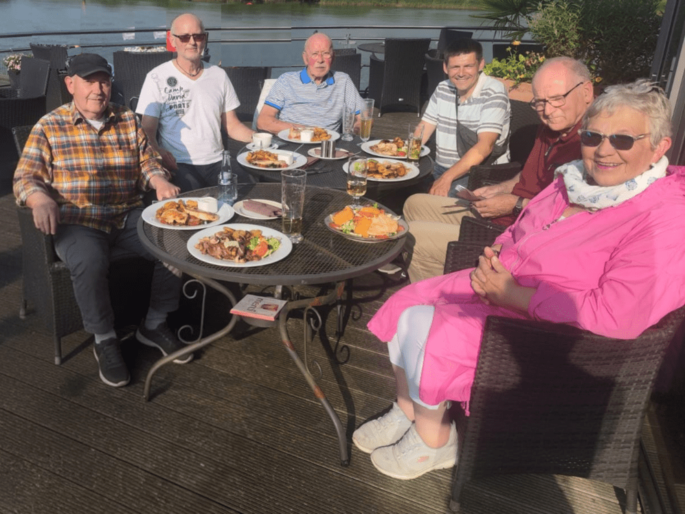 Sechs Personen sitzen an einem Tisch im Freien und genießen ein Mittagessen mit verschiedenen Gerichten.