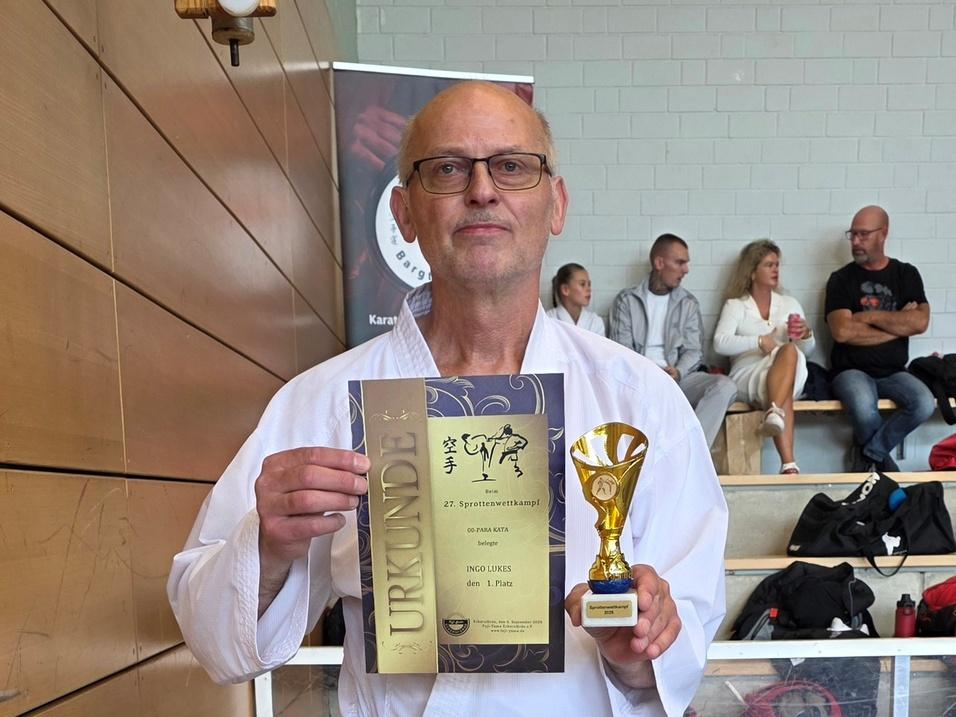 Ein Mann im weißen Karateanzug mit blauem Gürtel hält eine Urkunde und einen Pokal in der Hand und steht barfuß in einer Sporthalle vor einer Holzverkleidung.