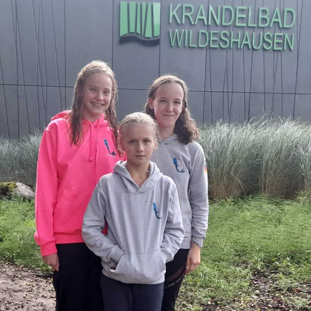 Drei M&auml;dchen in Freizeitkleidung stehen vor dem Krandelbad Wildeshausen, umgeben von Gras und Erde.