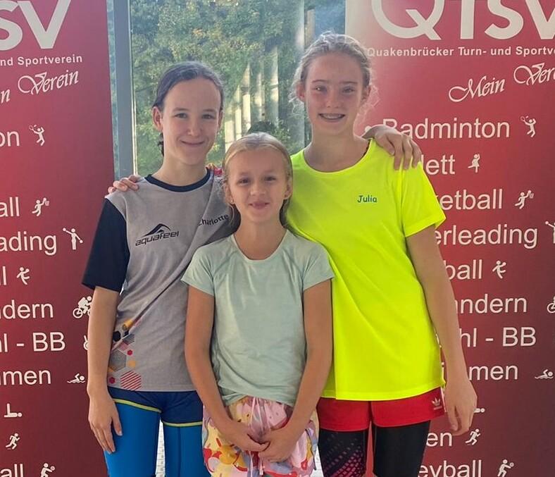 Drei M&auml;dchen stehen auf einem Siegertreppchen mit rot-wei&szlig;en QtSV-Postern im Hintergrund.