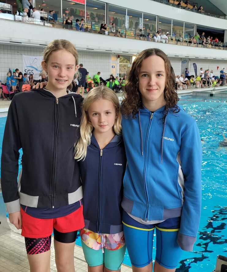 Drei M&auml;dchen in Bademode stehen am Beckenrand eines Schwimmbads, im Hintergrund sind Zuschauer und Wassersportler zu sehen.