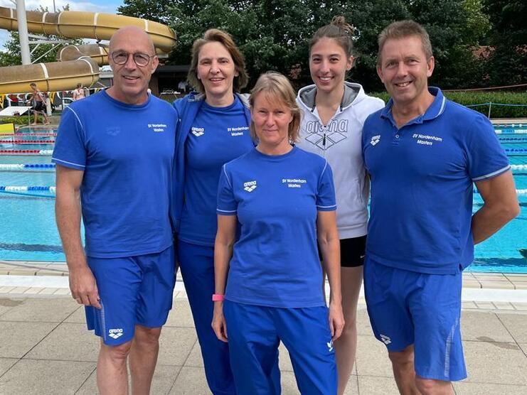 F&uuml;nf Personen in blauen Sportanz&uuml;gen stehen vor einem Schwimmbecken, umgeben von B&auml;umen und einem Wasserrutschenturm.