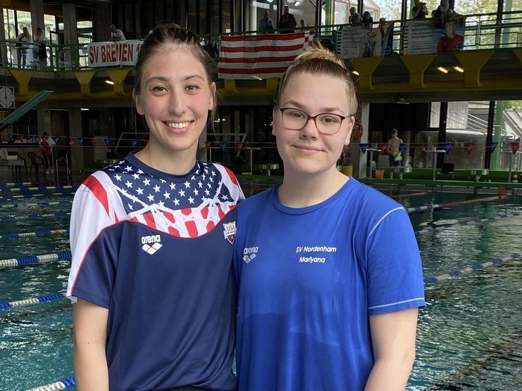 Zwei junge Schwimmerinnen stehen am Beckenrand eines Schwimmbades, umgeben von Wasser und Zuschauern.