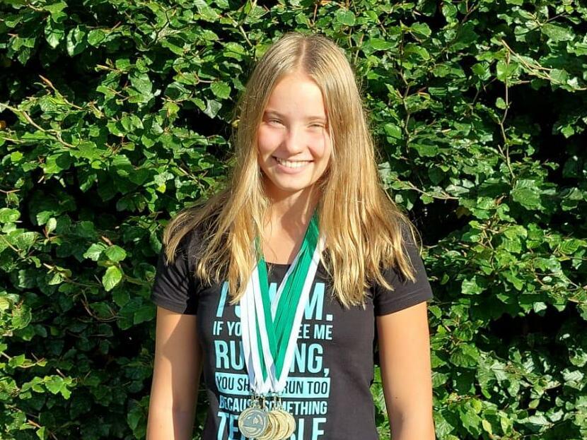 Junge mit langer blondem Haar und Medaille steht vor einer grünen Hecke, trägt ein schwarzes T-Shirt und Sportleggings.