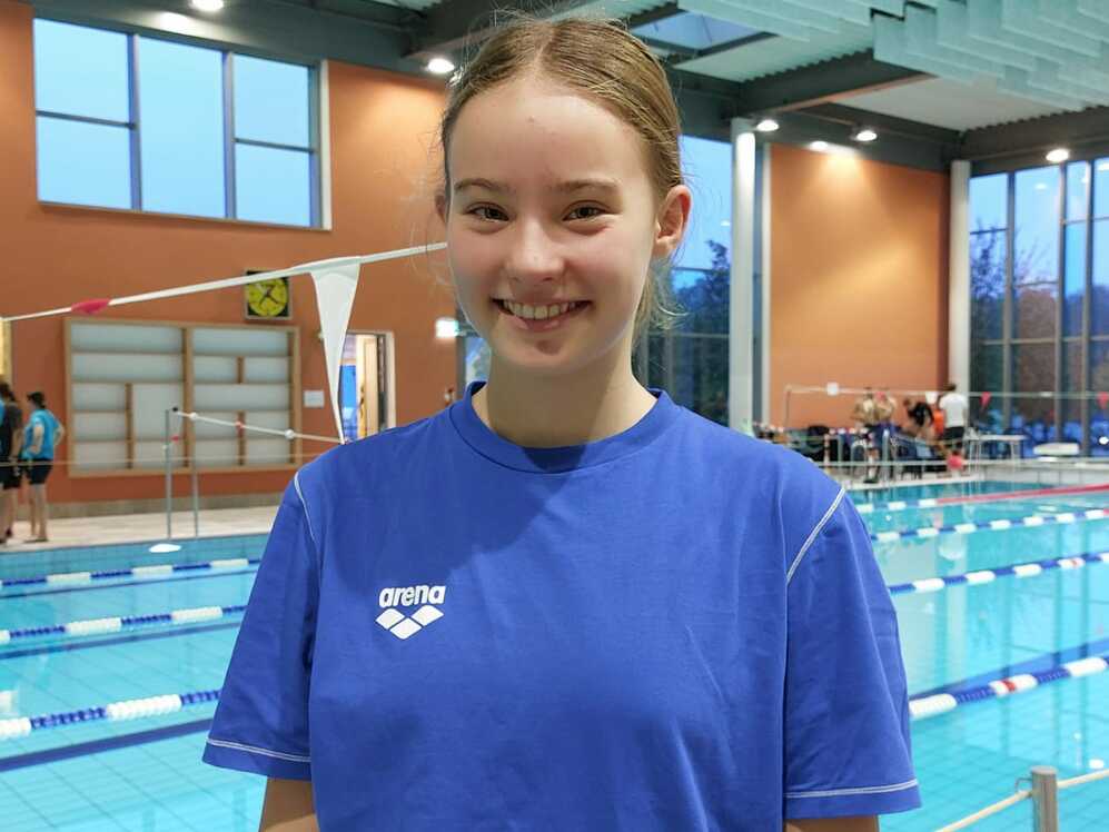 Lächelnde Schwimmerin in blauer T-Shirt vor einem Hallenbad mit Schwimmbecken und Sportlern im Hintergrund.