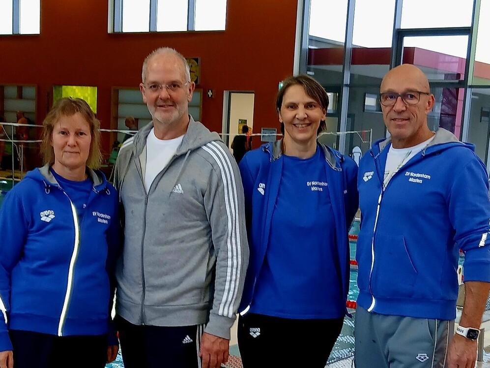 Schwimmer in blauen und grauen Jacken posieren vor einem Wettkampfbecken.