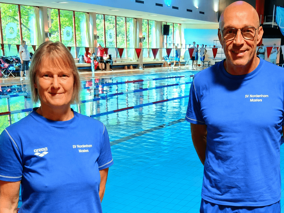 Zwei Personen in blauen T-Shirts mit dem Aufdruck 'SV Nordenham Masters' stehen am Beckenrand eines Hallenbads.