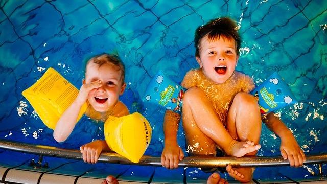 Zwei fr&ouml;hliche Kinder mit Schwimmhilfen im Wasser, l&auml;cheln und halten sich am Rand des Schwimmbeckens fest.