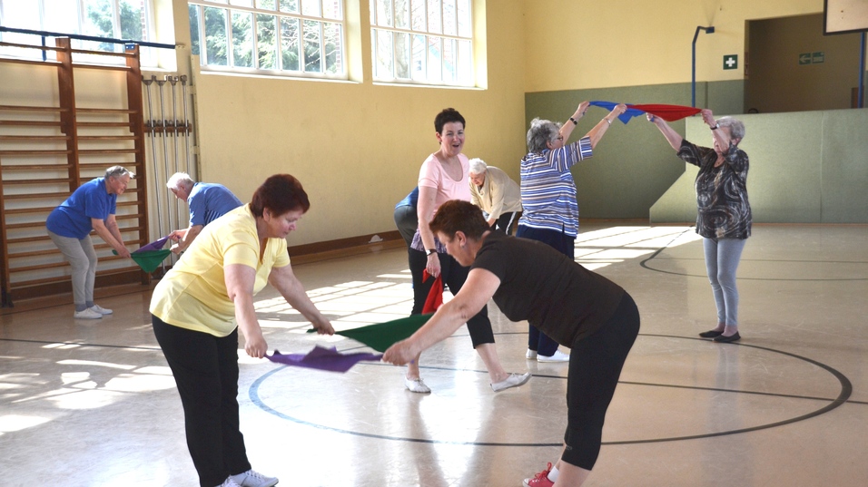 Gruppe von Senioren führt Bewegungsübungen mit bunten Tüchern in einem hellen Gymnastikraum durch.