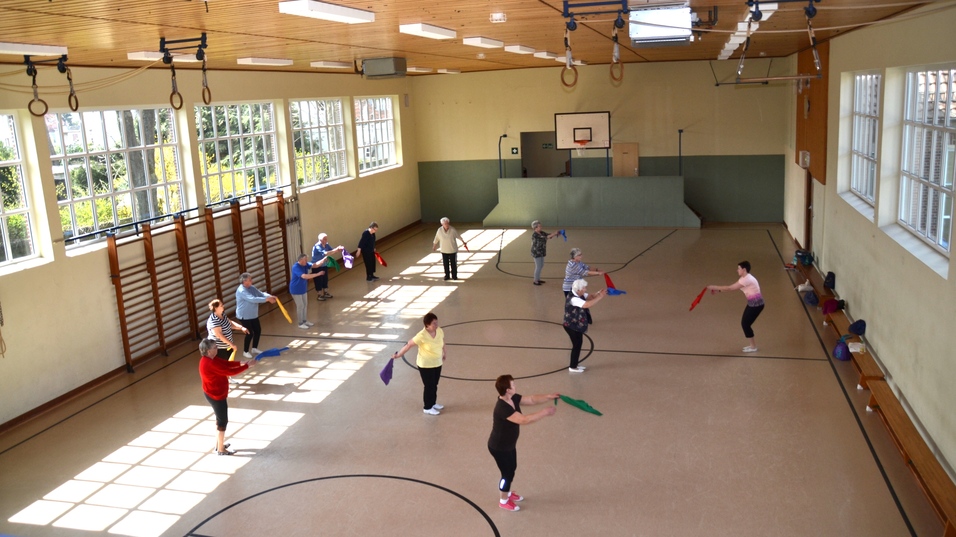 Gruppe älterer Menschen macht Gymnastik mit bunten Tüchern in einer hellen Turnhalle.