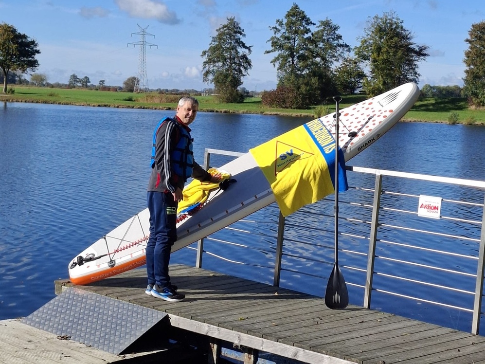 Lächelnder Mann mit einem SUP-Board in der Hand, vor einem See stehend.