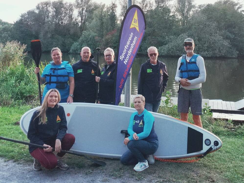 Gruppe von sieben Personen mit Paddelboards und Paddeln an einem Flussufer bei Tageslicht.