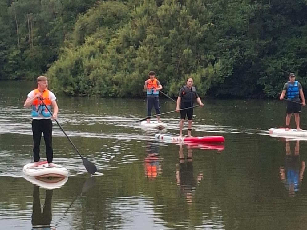 Mehrere Personen stehen auf SUP-Boards auf einem ruhigen See vor dicht bewachsenem Ufer.
