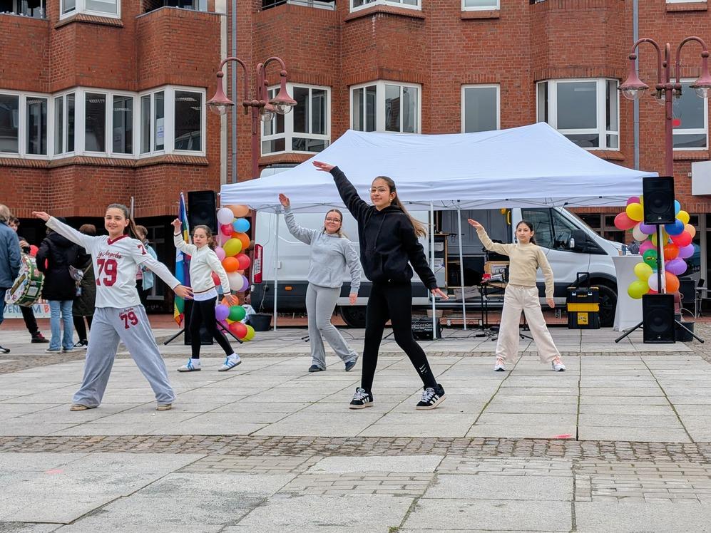 F&uuml;nf junge Frauen tanzen auf einem gepflasterten Platz vor einem wei&szlig;en Zelt und einem roten Backsteingeb&auml;ude.